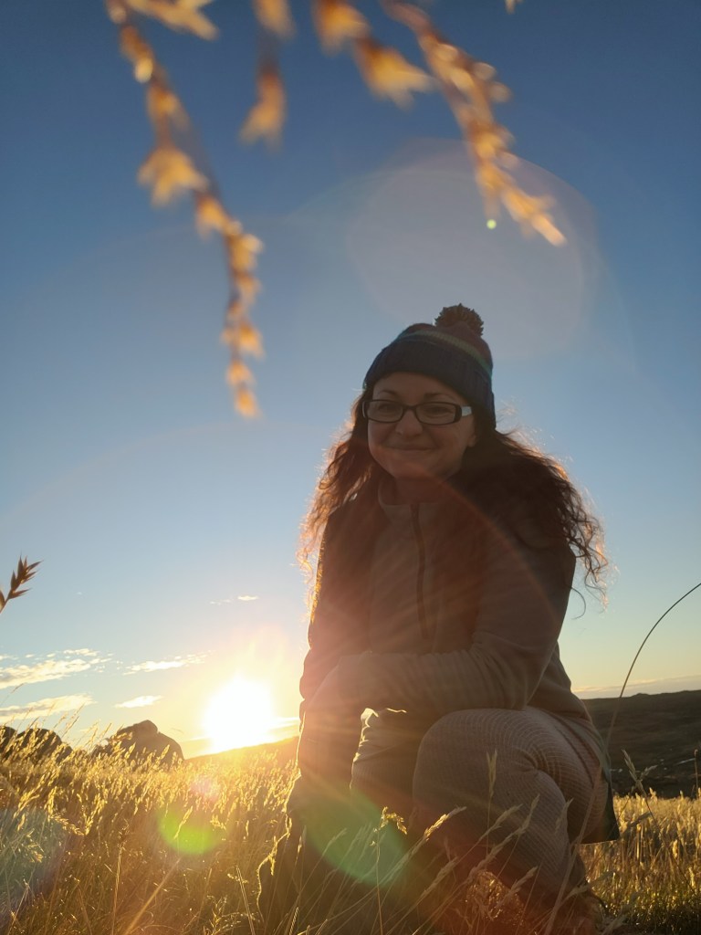 A woman kneeling in a field at sunrise, wearing a blue beanie and smiling with a scenic backdrop.